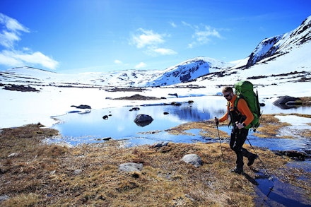 Man with rucksack and trekking poles walking by lake in snow covered mountain landscape Man with rucksack and trekking poles walking by lake in snow covered mountain landscape