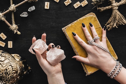 top view of witch holding crystals and touching aged book of dark magic near voodoo doll and runes on black; Shutterstock ID 1763528189; purchase_order: -; job: -; client: -; other: - Hands holding crystals with runes and old book