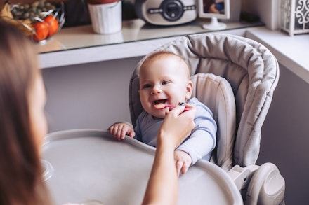 Mom feeds the baby from the spoon in the kitchen. A child is sitting in a child's chair. baby in a highchair being fed by mum