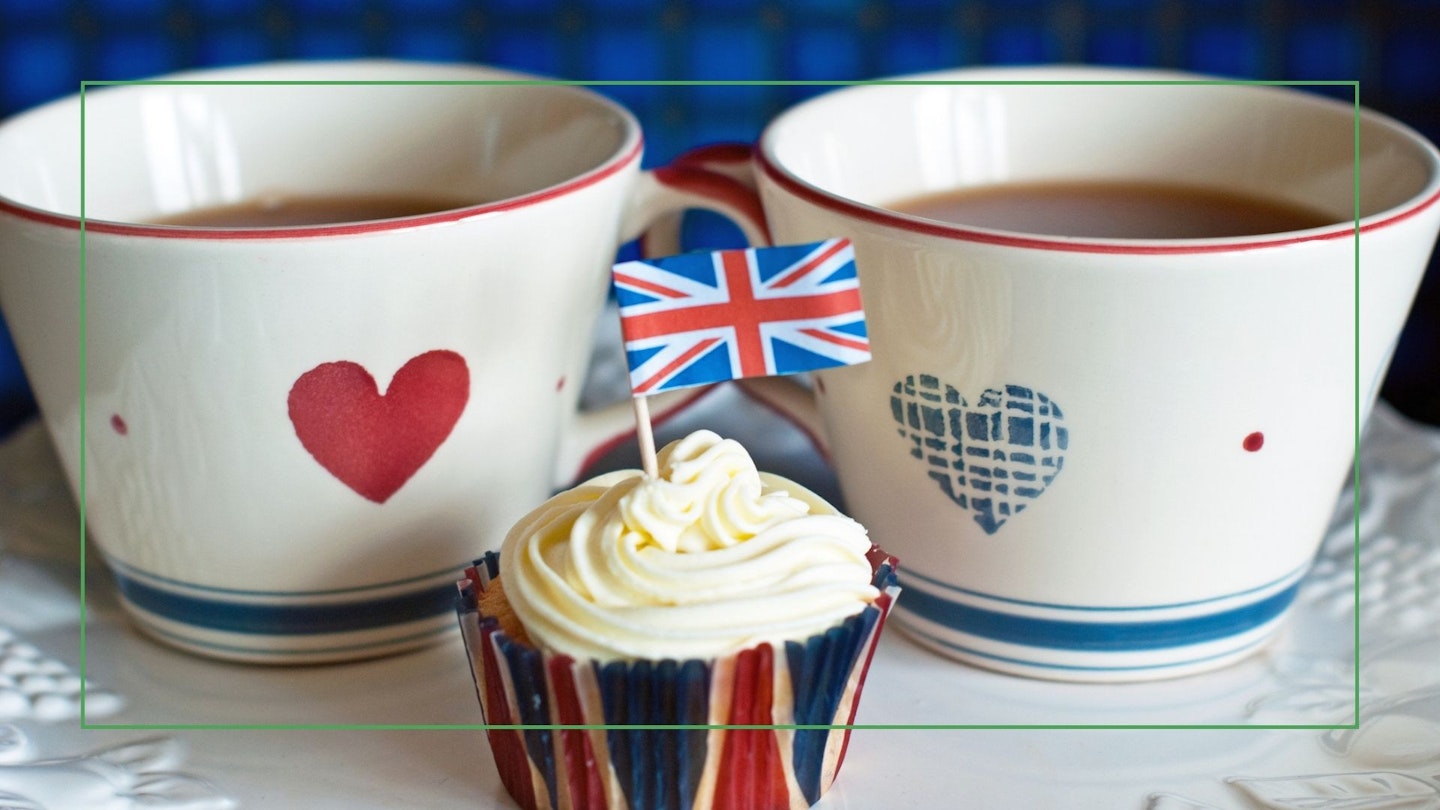 Two tea/coffee cups decorated with a red heart on one and a blue heart on the other. In front stands a cupcake in a Union Jack case with white icing swirled on top and decorated with a mini-Union Jack flag.