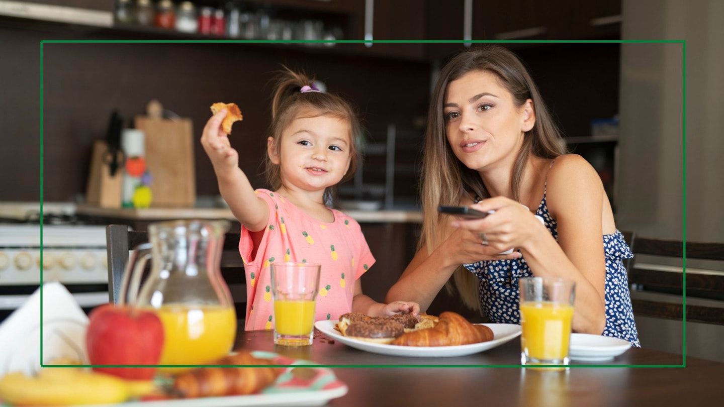 A mum and young daughter eat breakfast in the kitchen and watch TV