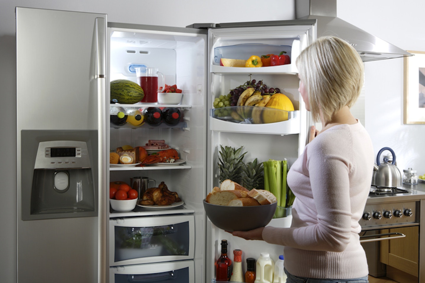 A young woman looking in an open fridge filled with food and drinks in a home kitchen