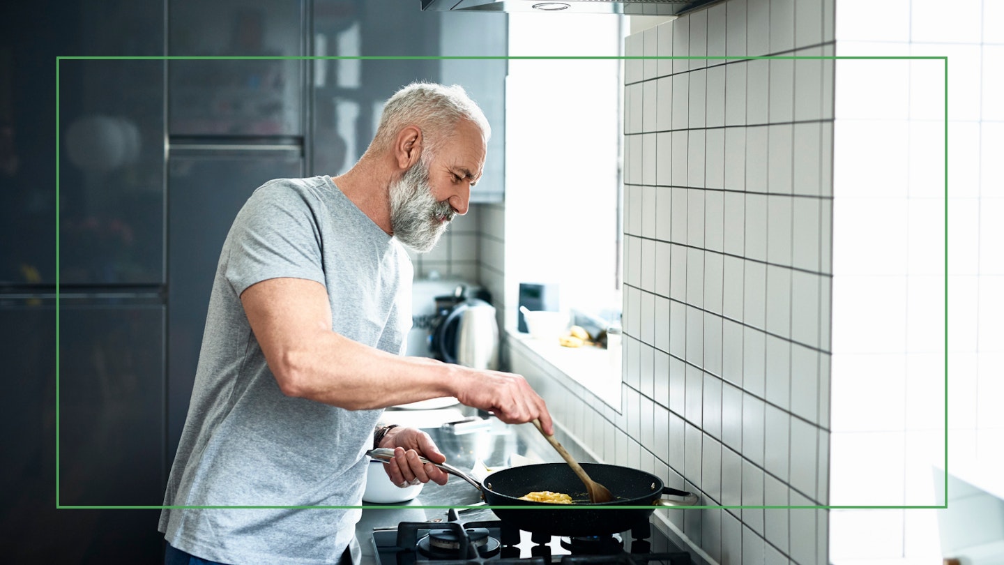 Side view portrait of man in his 50s cooking dinner, standing by stove and cooking on a gas hob, white tiled wall with natural light