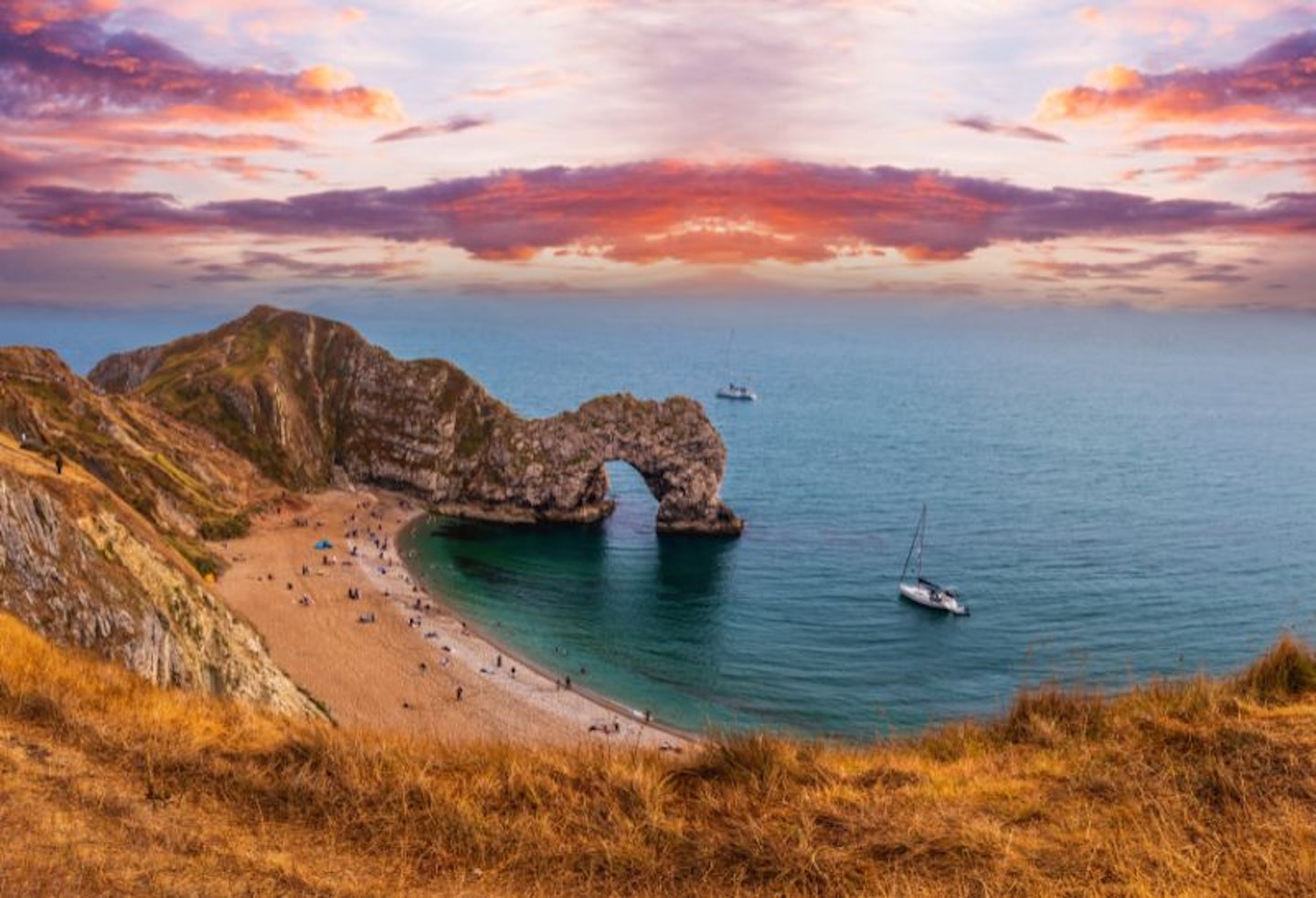 Durdle Door in Dorset at sunset, with a beach and the rock with a hole through and a boat on the water inDoreset