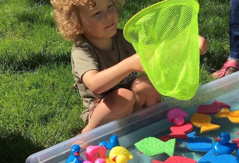 A little boy using a green fishing net to catch sponge fishes from a bucket of water