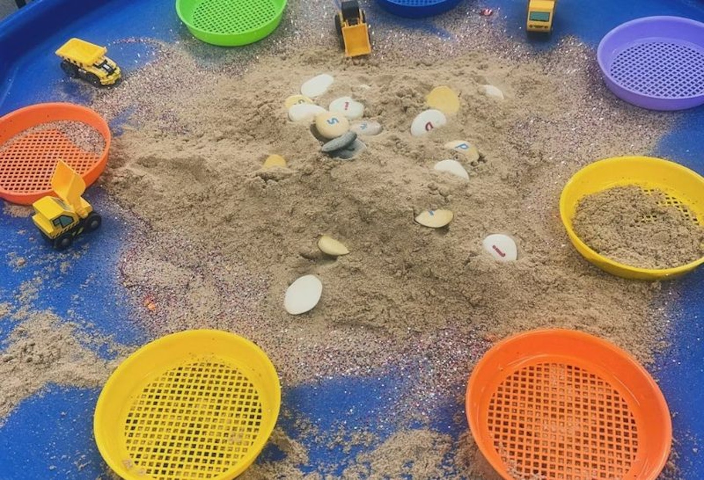 Colourful play sieves on a blue tuff table with sand as an activity for one year olds