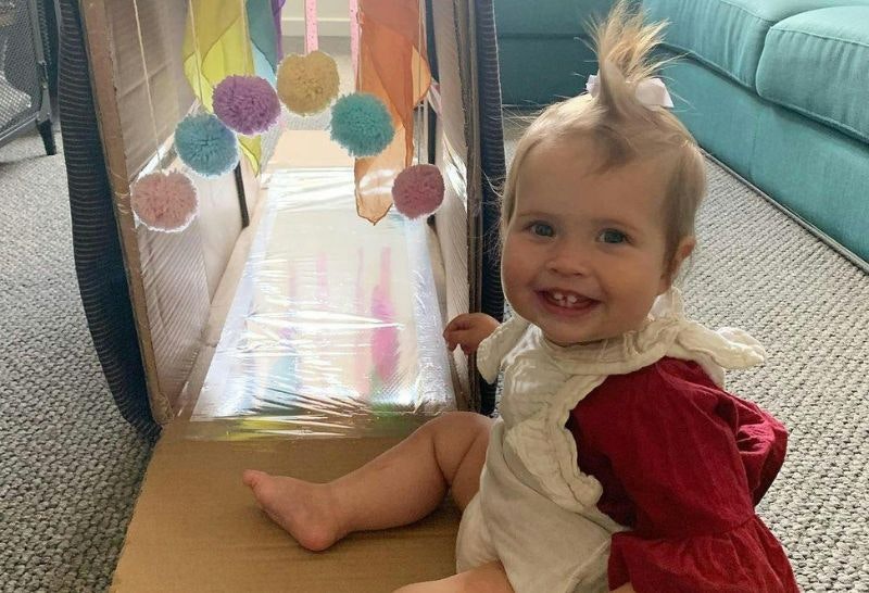 A one year old sitting in front of a cardboard box which is open with silver foil inside and colourful balls hung from the top