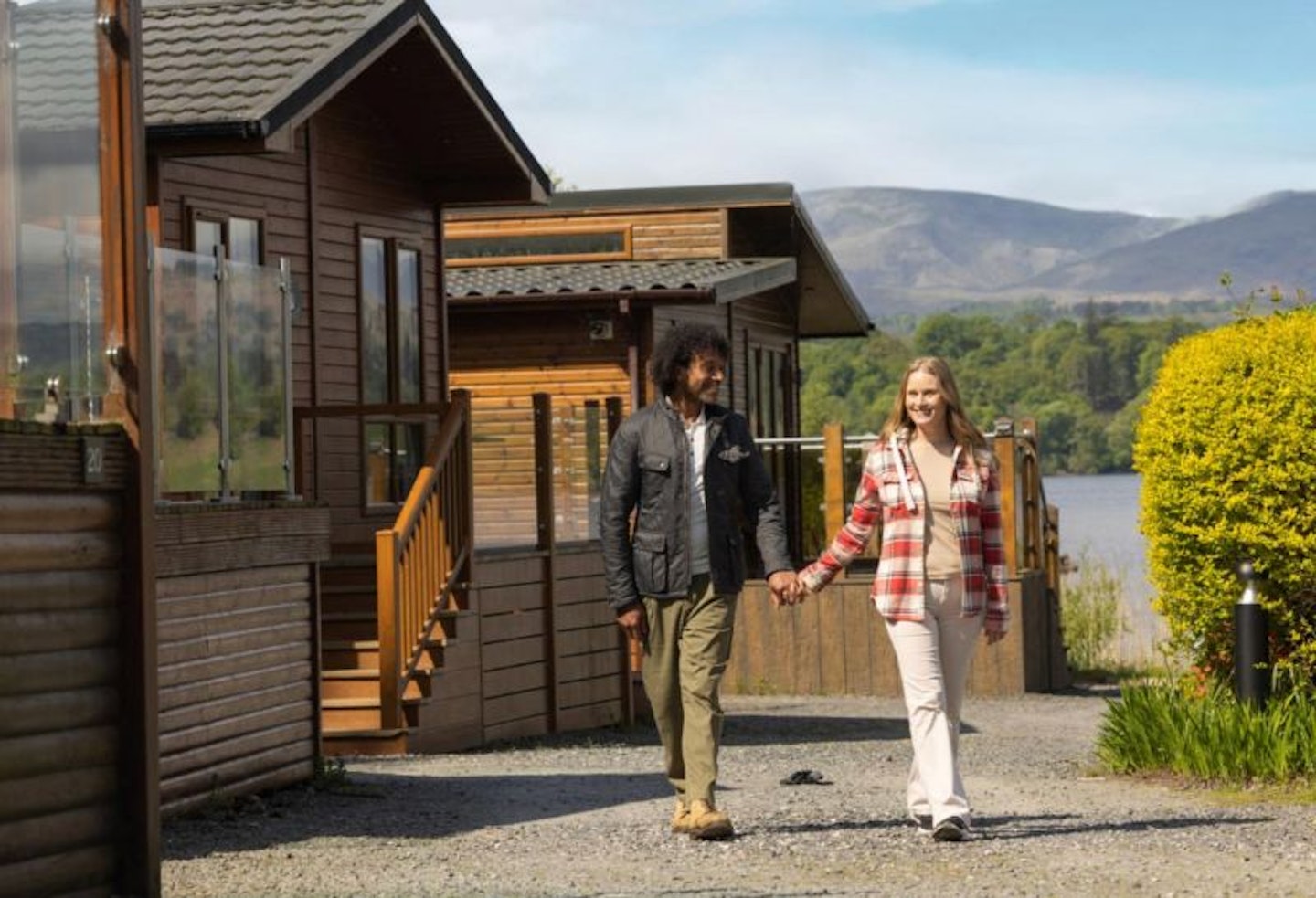 A couple walking past two holiday lodges on the edge of a lake with hills in the background at the White Cross Bay Parkdean Holiday Park