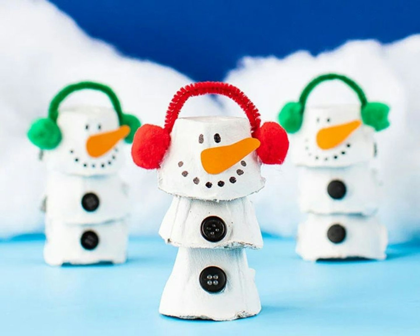 Three snowmen made out of egg cartons with ear muffs on a snowy backdrop
