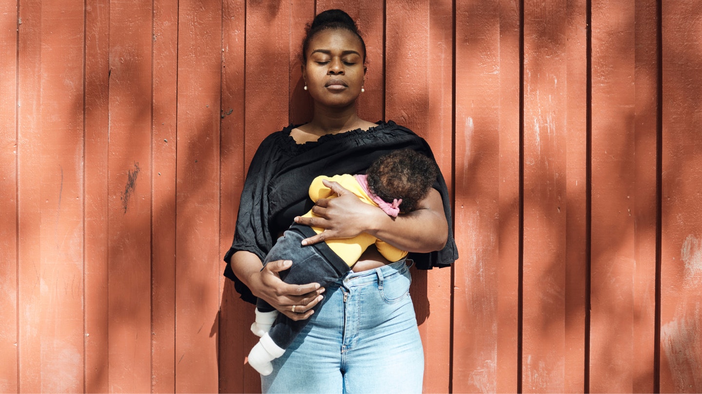 an with eyes closed breastfeeding toddler daughter in front of wall on sunny day