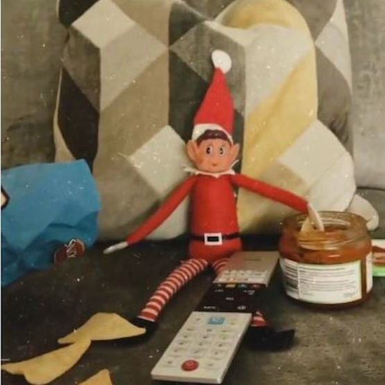 An Elf on the Shelf sitting on the sofa with chips and dip and the TV remote