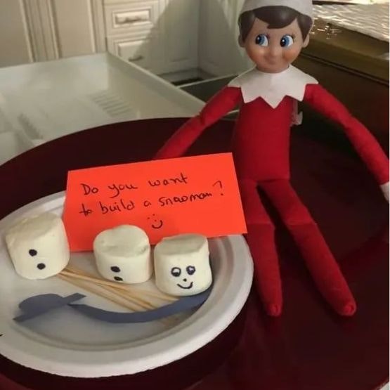 An elf with a sign saying do you wnt to build a snowman with 3 white marshmallows with face and buttons on