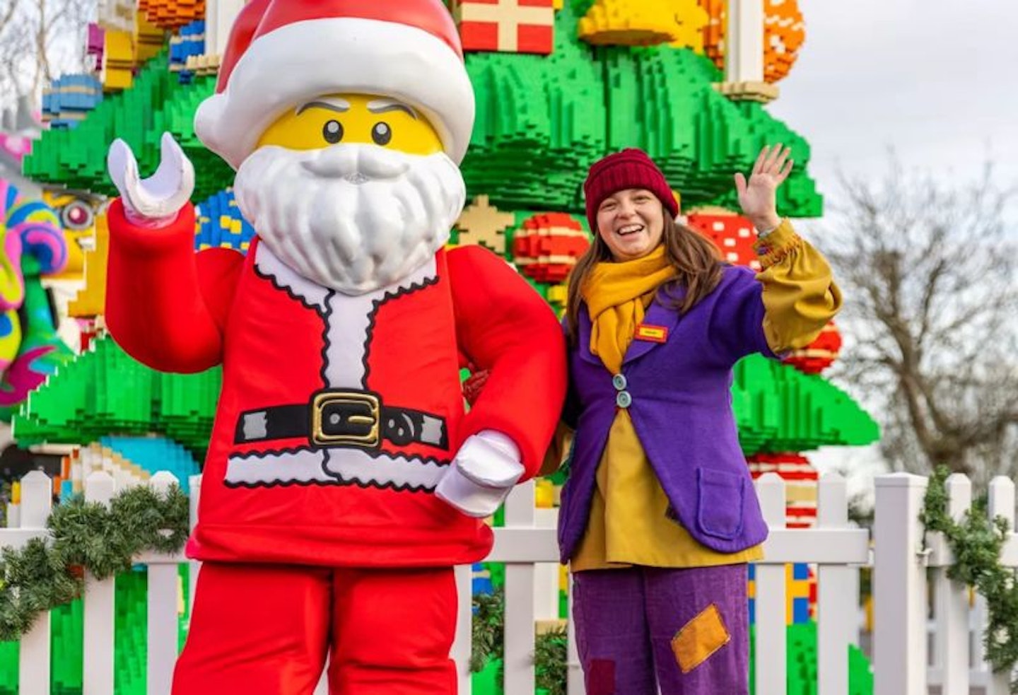 A lego Father Christmas and woman standing in front of a Duplo Christmas tree and waving at the camera