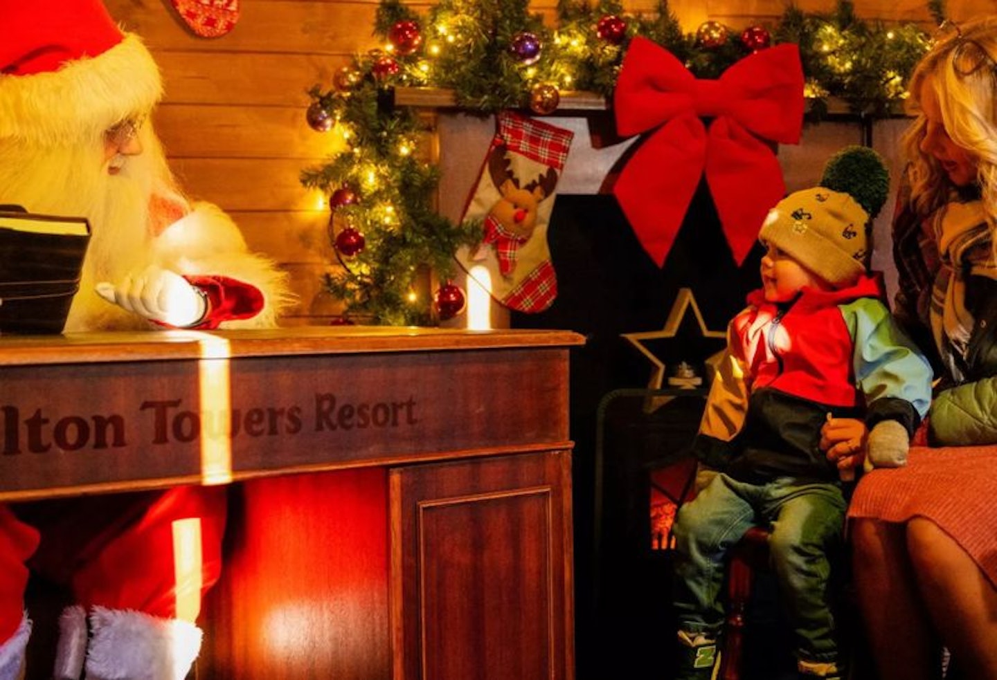 A little boy sat on his mum's lap meeting Santa who's sat behind a desk that says 'Alton Towers Resort' on.