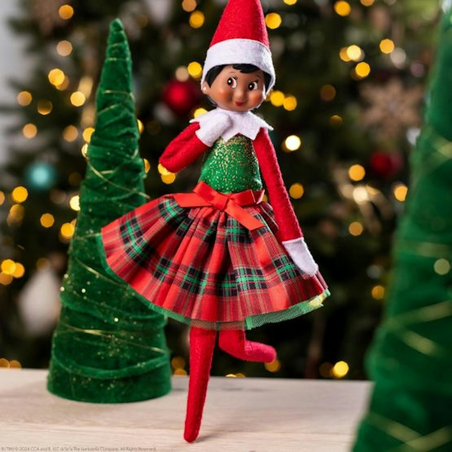 An Elf on the Shelf wearing a tartan skirt and green top in front of a Christmas tree