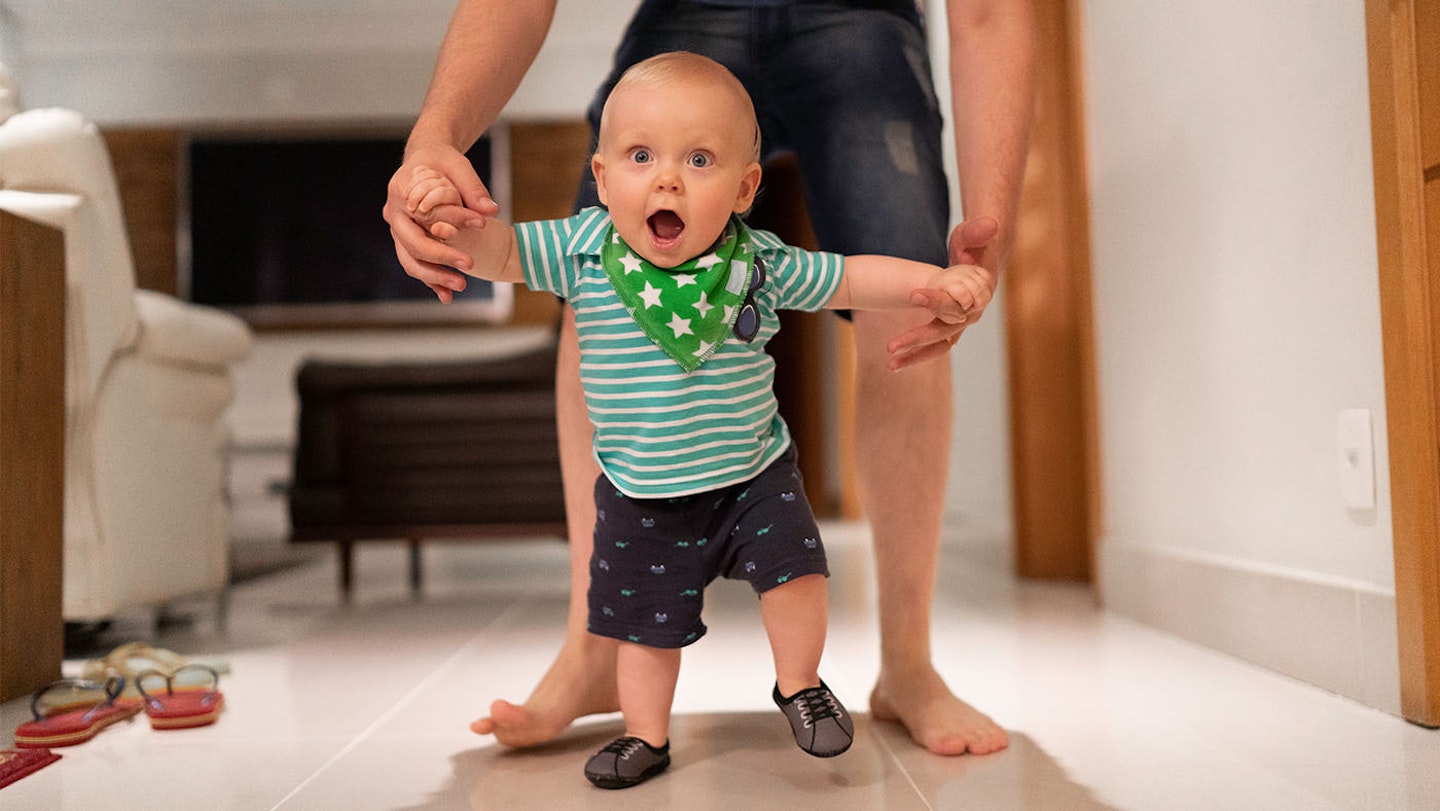 A little boy is looking directly at the camera with a shocked expression, while an adult assists him by holding his arms, helping him to walk.