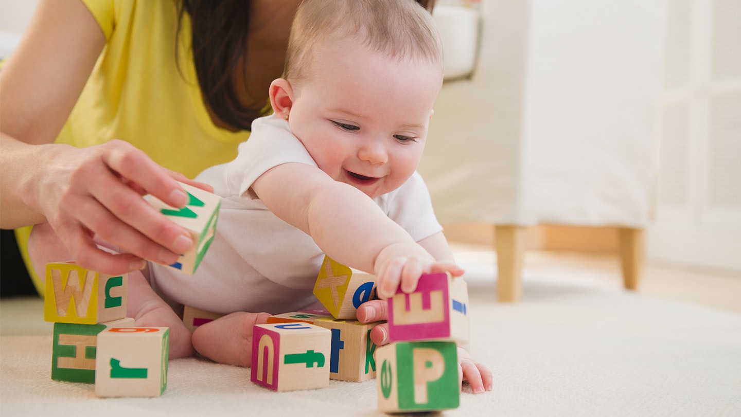 A little baby enjoying playing with some building blocks with their guadian in the background.