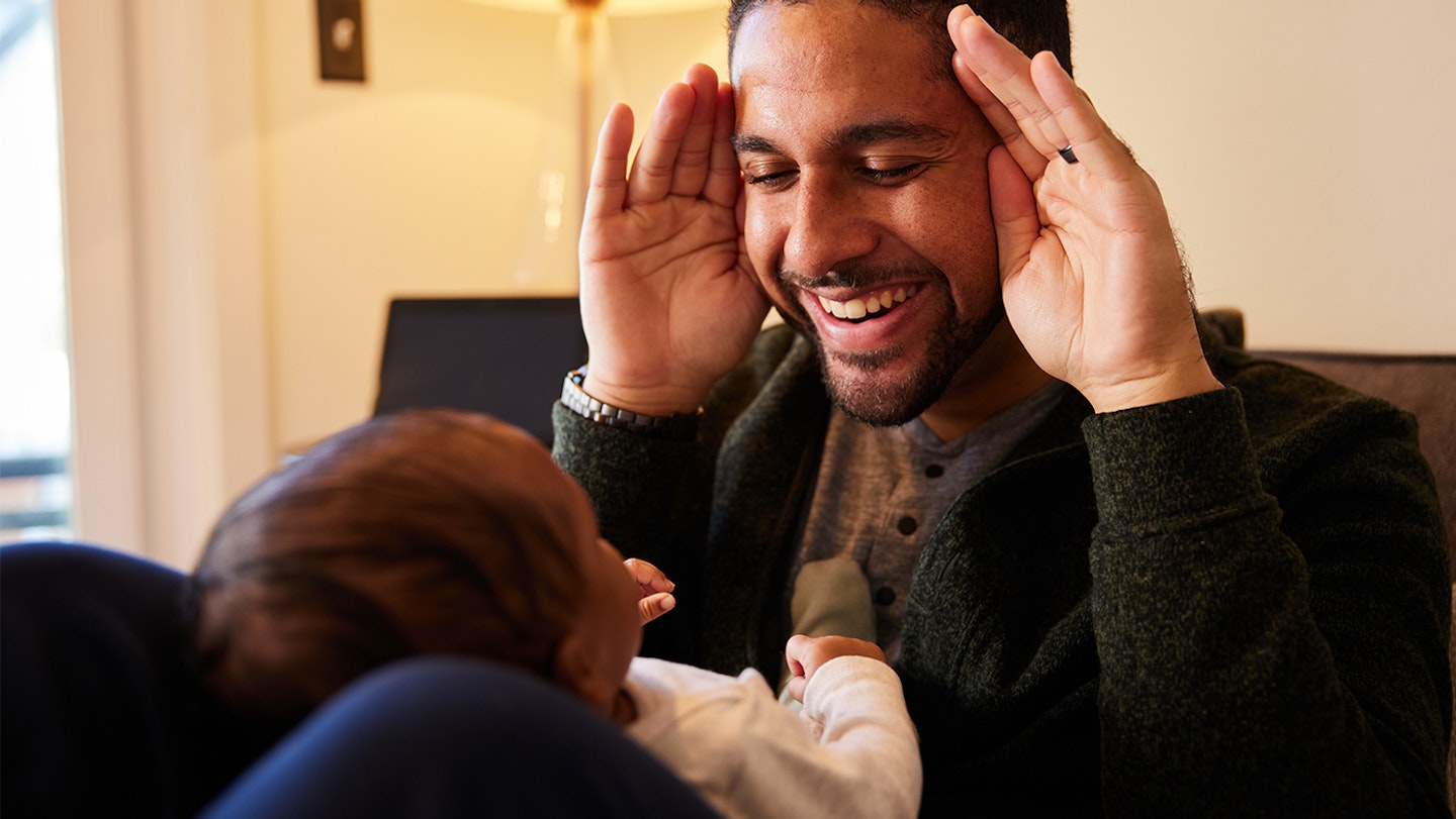 A father and his baby playing peekaboo. The father is happy and trying to encourage the baby to smile.