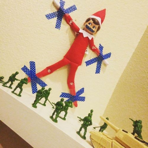 An elf taped to the wall with toy soldiers in front