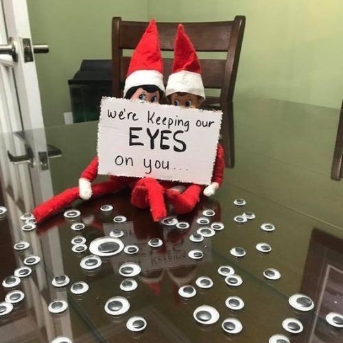 Two elf on the shelf with decorative eyes spread around the table and a sign saying 
