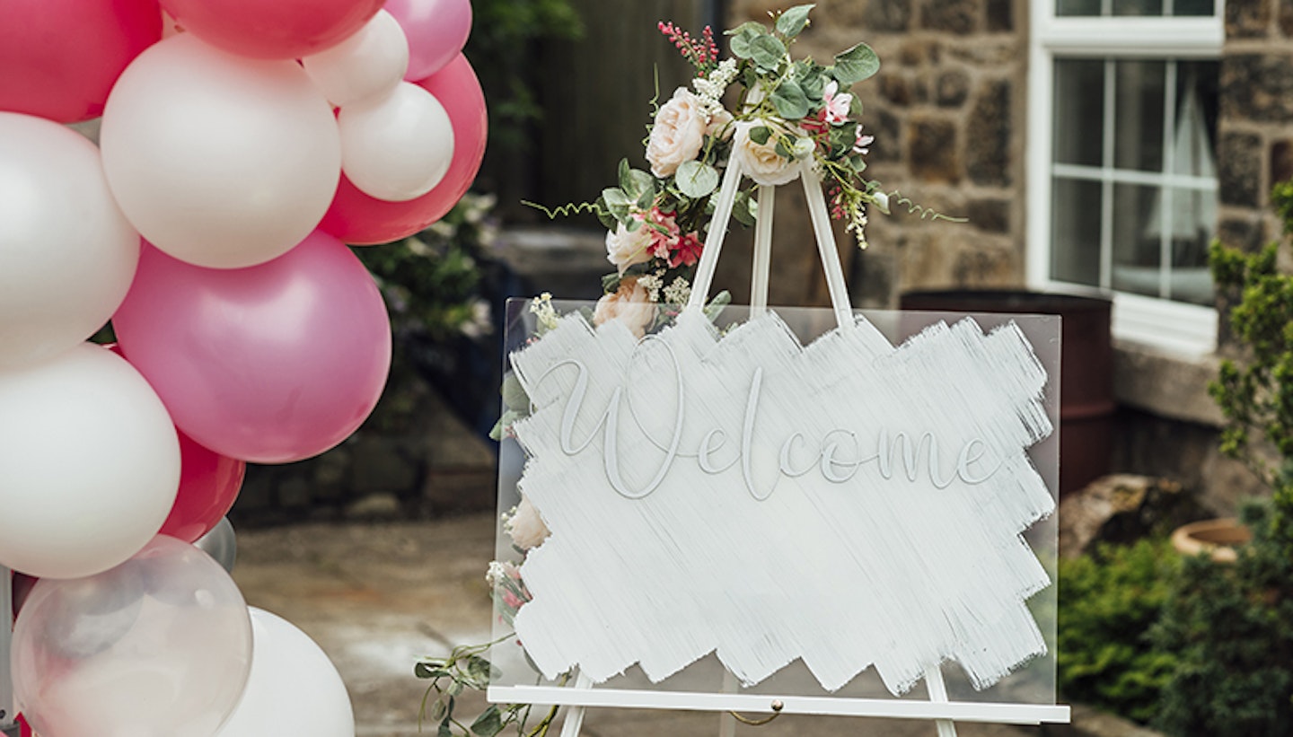A shot of a welcome sign in a garden next to decorative balloons on a summers day.