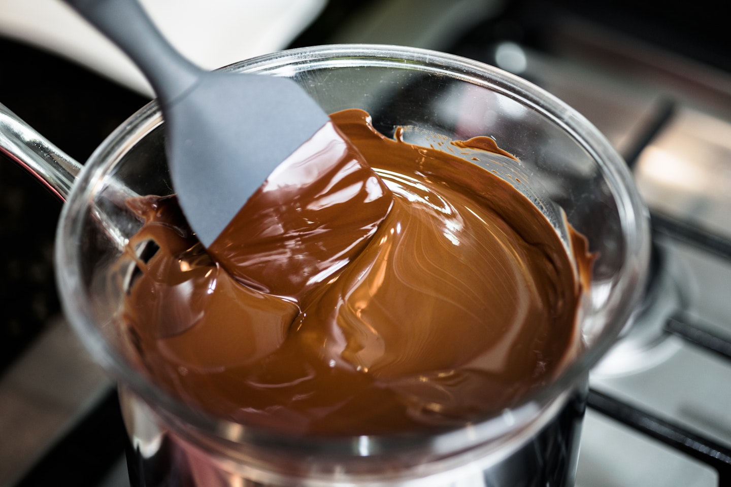 Close-up view of mixing chocolate while melting in transparent glass dish. Preparing dessert in home kitchen. Warming up ingredient on gas stove.