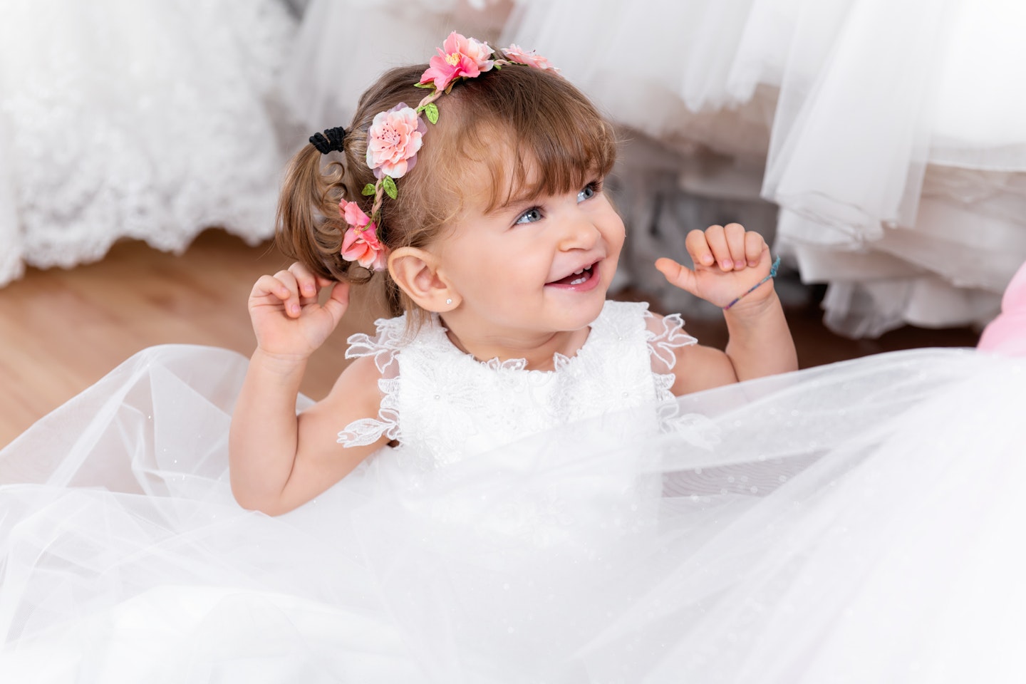 Portrait of 18 months old baby girl smiling and playing at store. Small girl wearing flower wreath and trying on dress at bridal boutique.