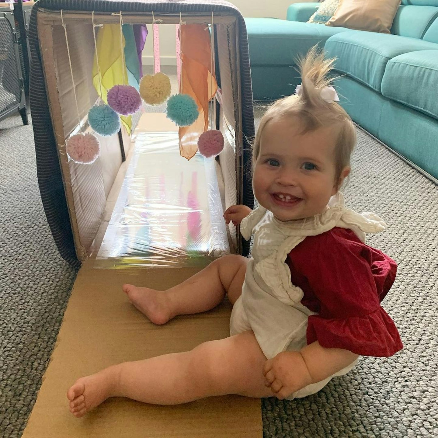 Toddler sitting in front of a cardboaqrd tunnel made for sensory play with hanging pompoms