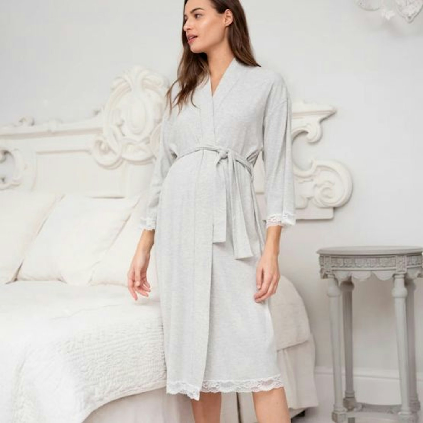 A woman wearing a grey maternity dressing gown in a bedroom with a white bed behind