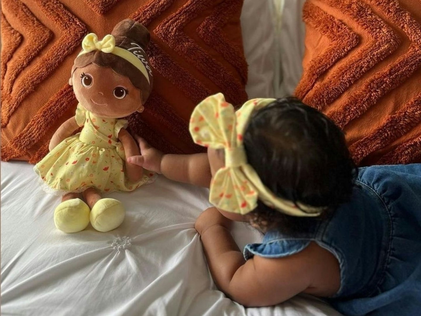 A toddler in a blue dress plays and a yellow headband plays with a Bibinee Doll in a matching dress