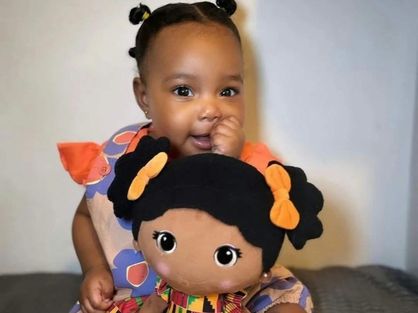 Toddler in brightly coloured clothes sits cuddling a Bibinee Doll in a Kente Print dress