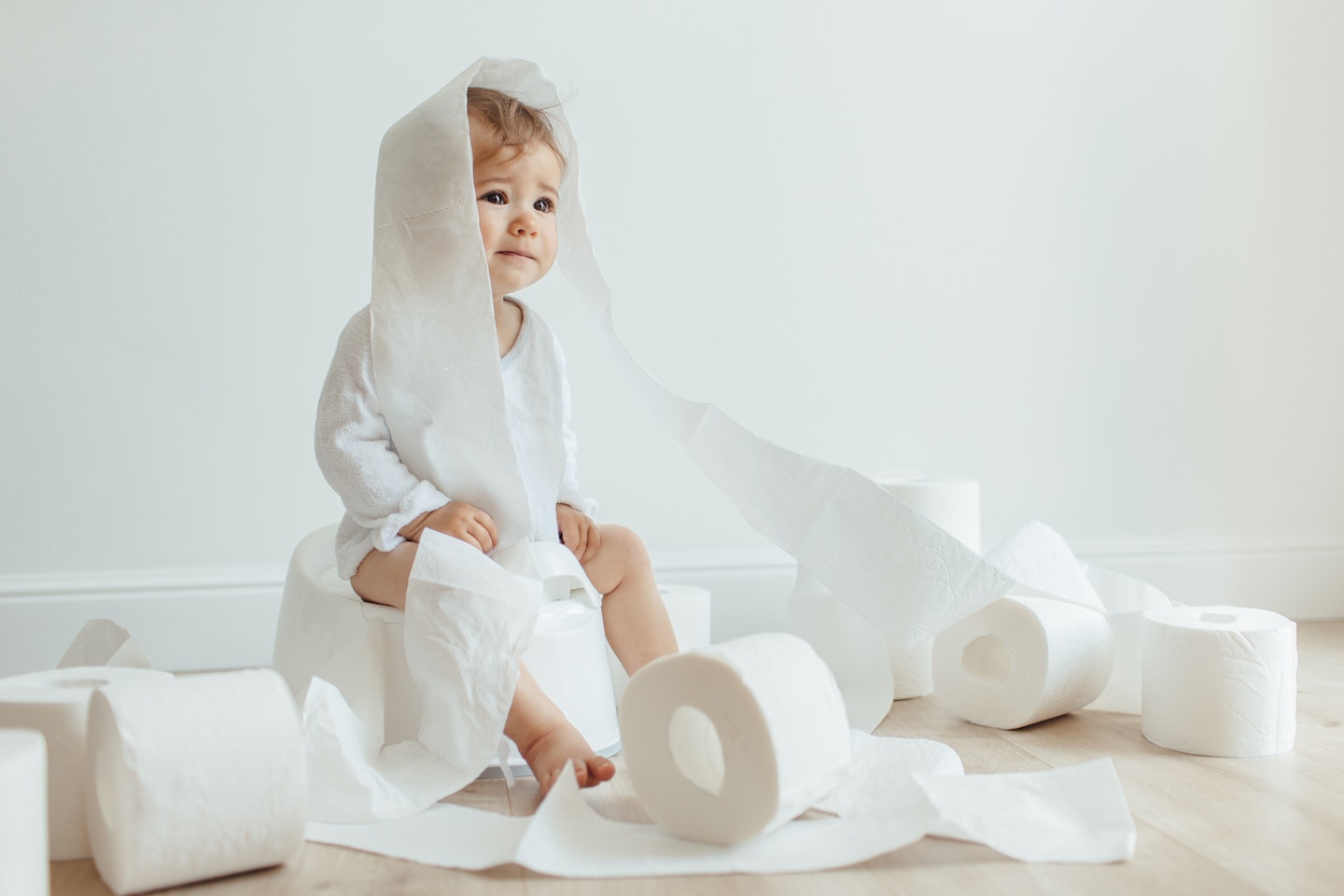 Cute baby girl sitting on white chamber pot with toilet paper rolls. Funny toddler sitting on potty chair and playing with toilet paper.