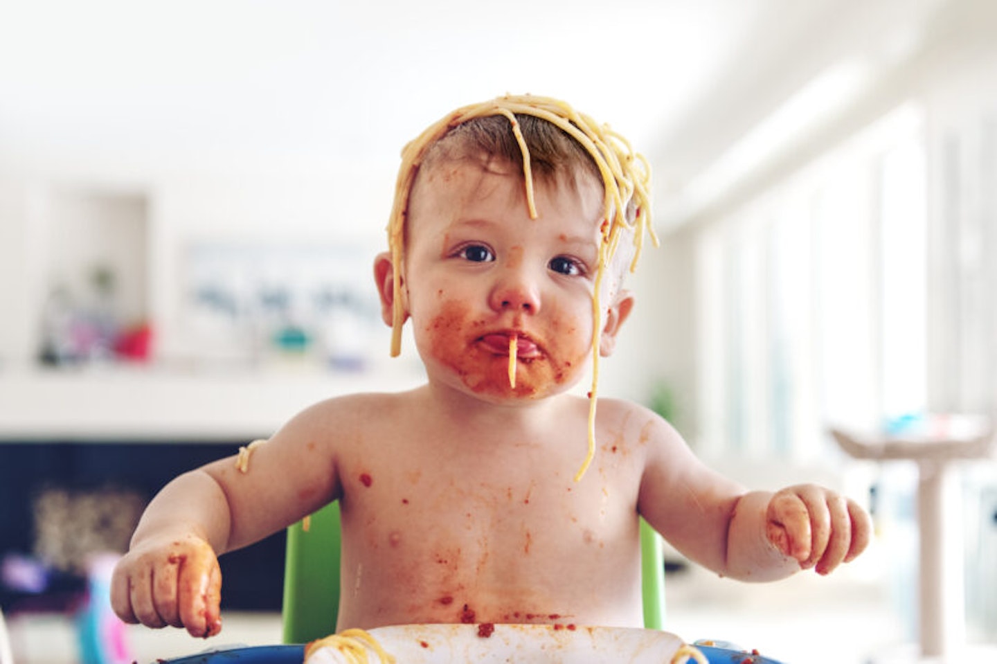 A baby sitting in a high chair with spaghetti on his head