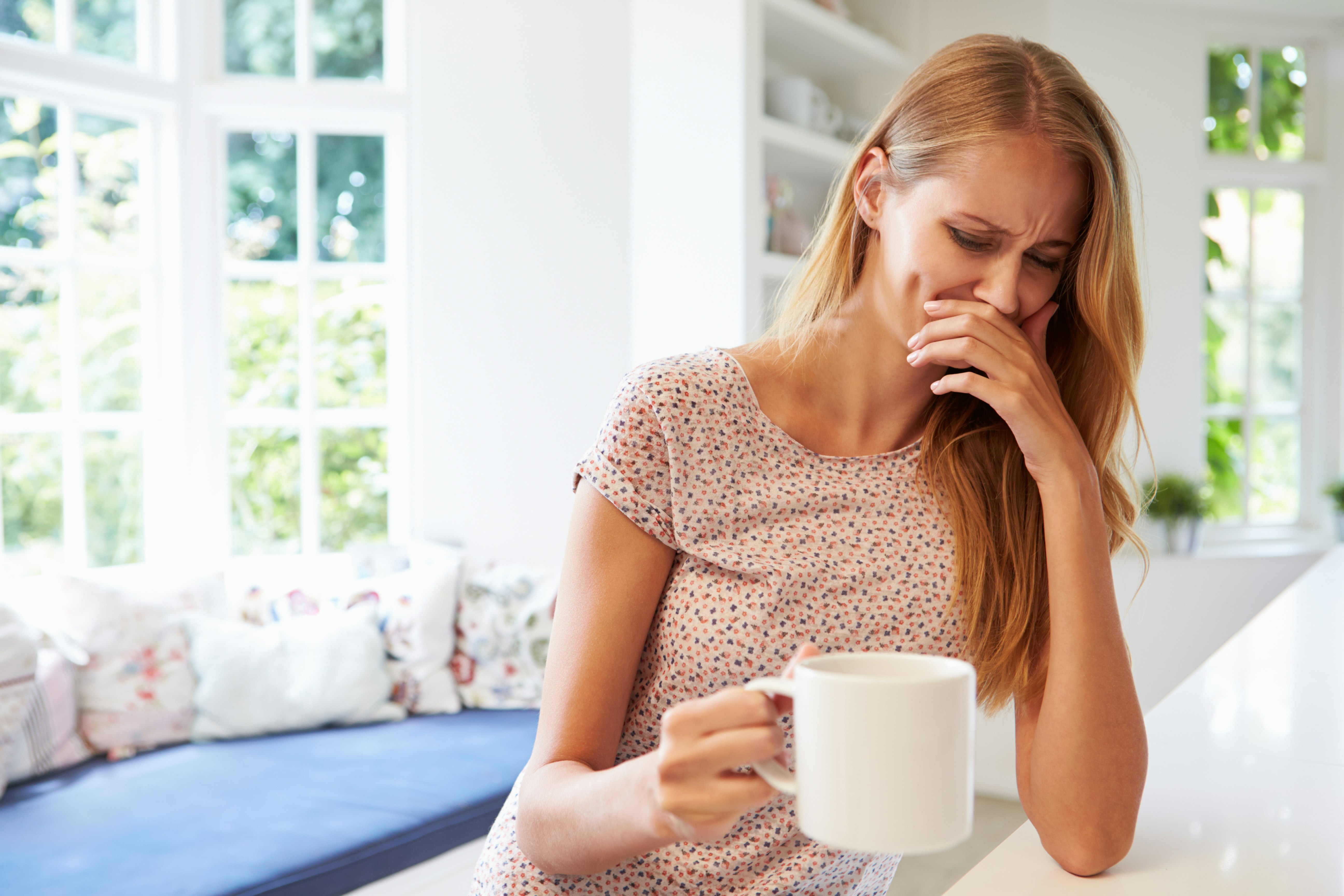 A woman sitting at a kitchen counter holding a mug and looking nauseous