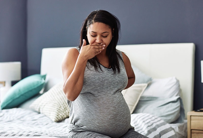 A pregnant woman sitting on the side of the bed with her hand to her mouth in a sign of feeling morning sickness