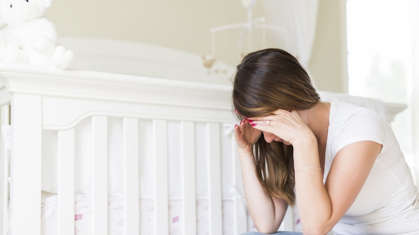 woman sits next to baby cot with head in hands feeling depressed and anxious