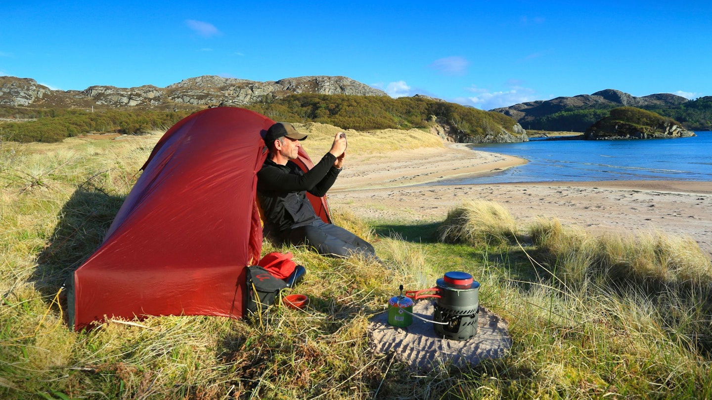 Hiker sitting outside tent taking a photo, Gruinard Bay, Scotland