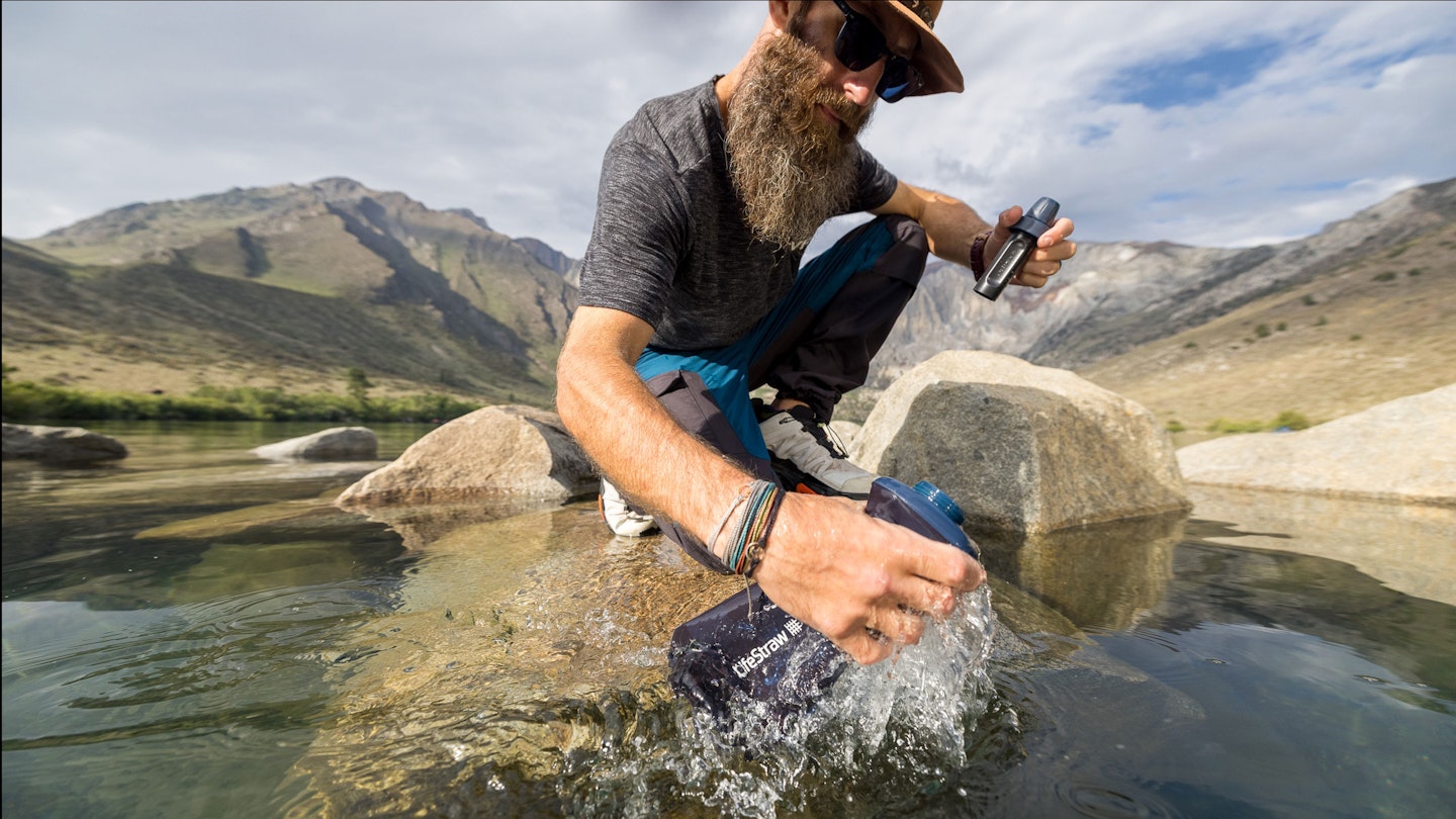 Bearded man in hat and sunglasses collecting water from a lake with a LifeStraw pouch and filter