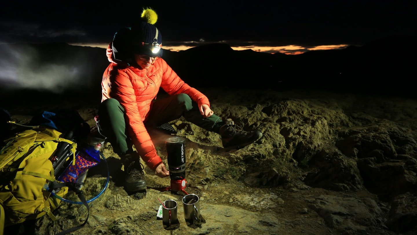 Hiker boiling a backpacking stove on a mountain with a headtorch