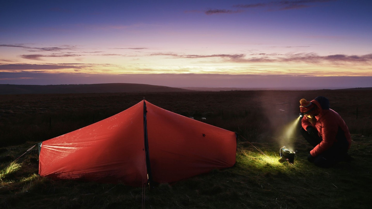 Hiker wearing ahead torch next to their tent using a stove at dusk