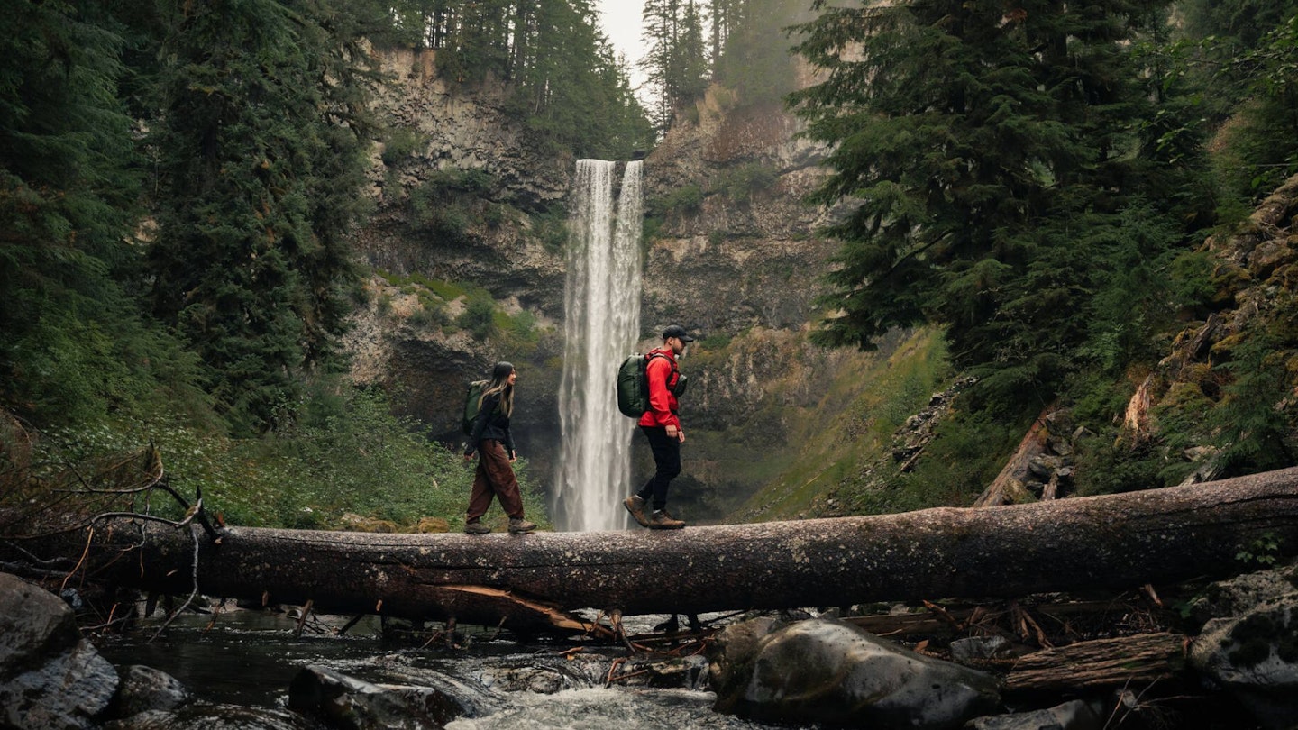 Hikers crossing a log near a waterfall wearing Osprey backpacks
