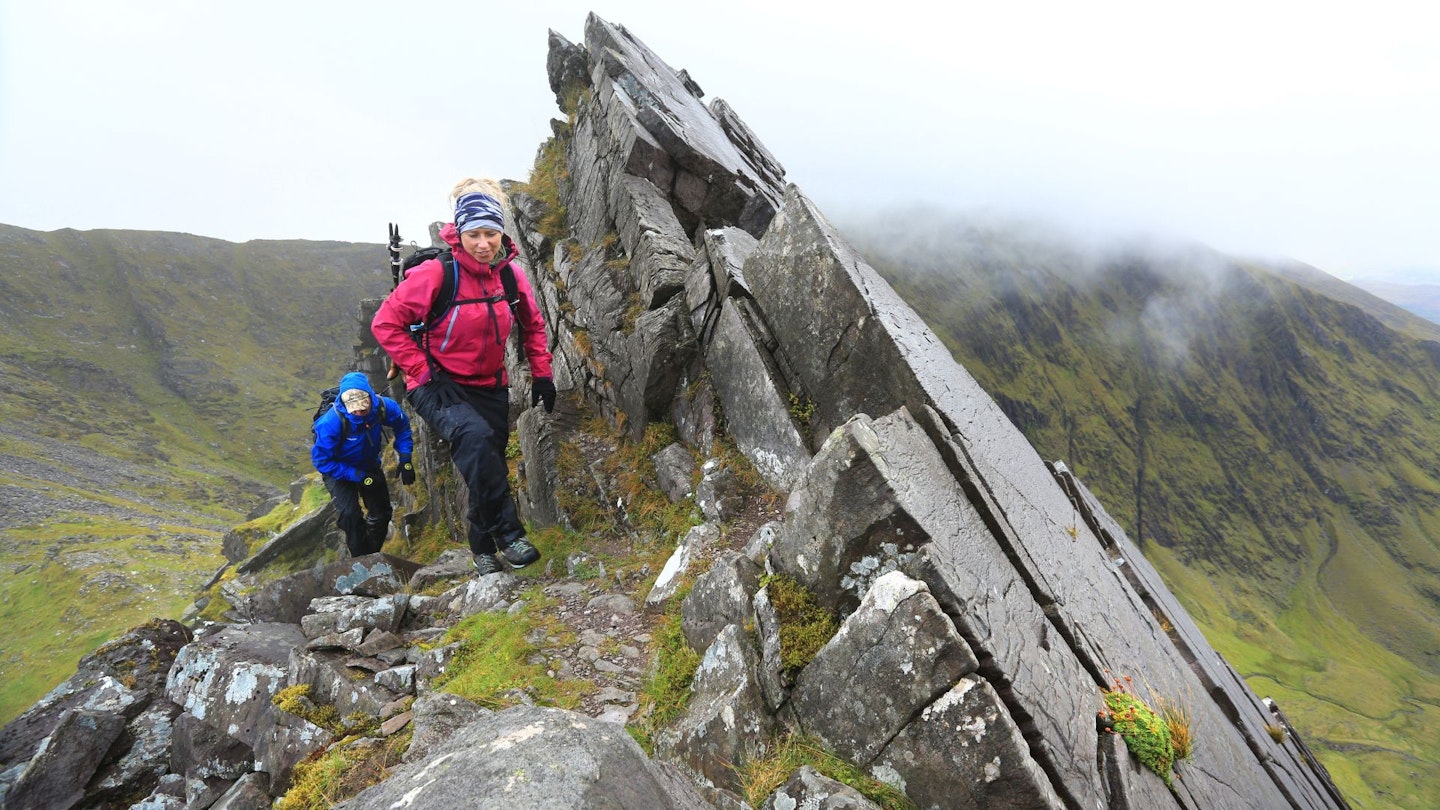 a woman and a man walk up a jagged ridge wearing waterproofs