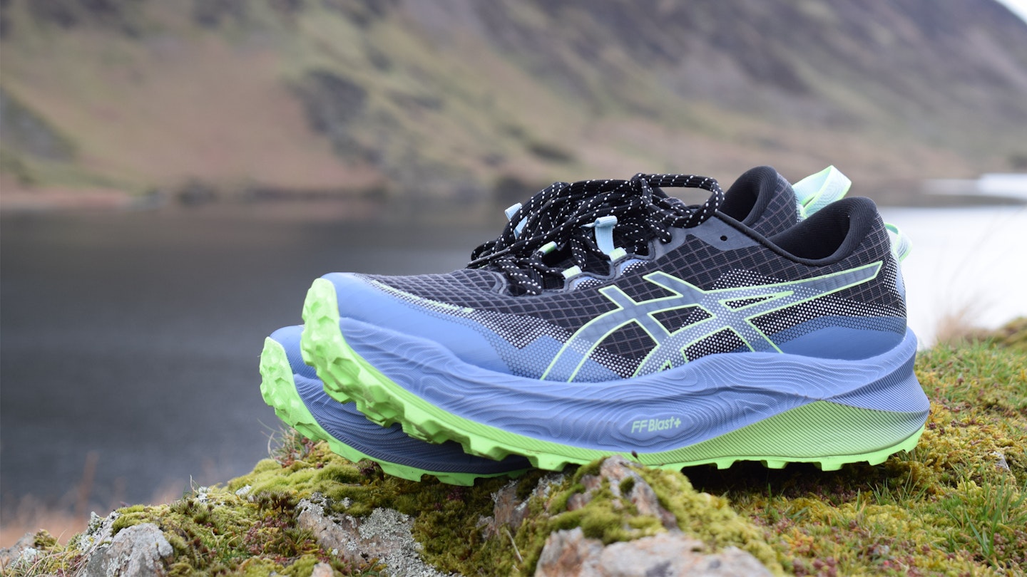 Asics Trabuco Max 3 trail running shoes on mossy rocks in front of a lake