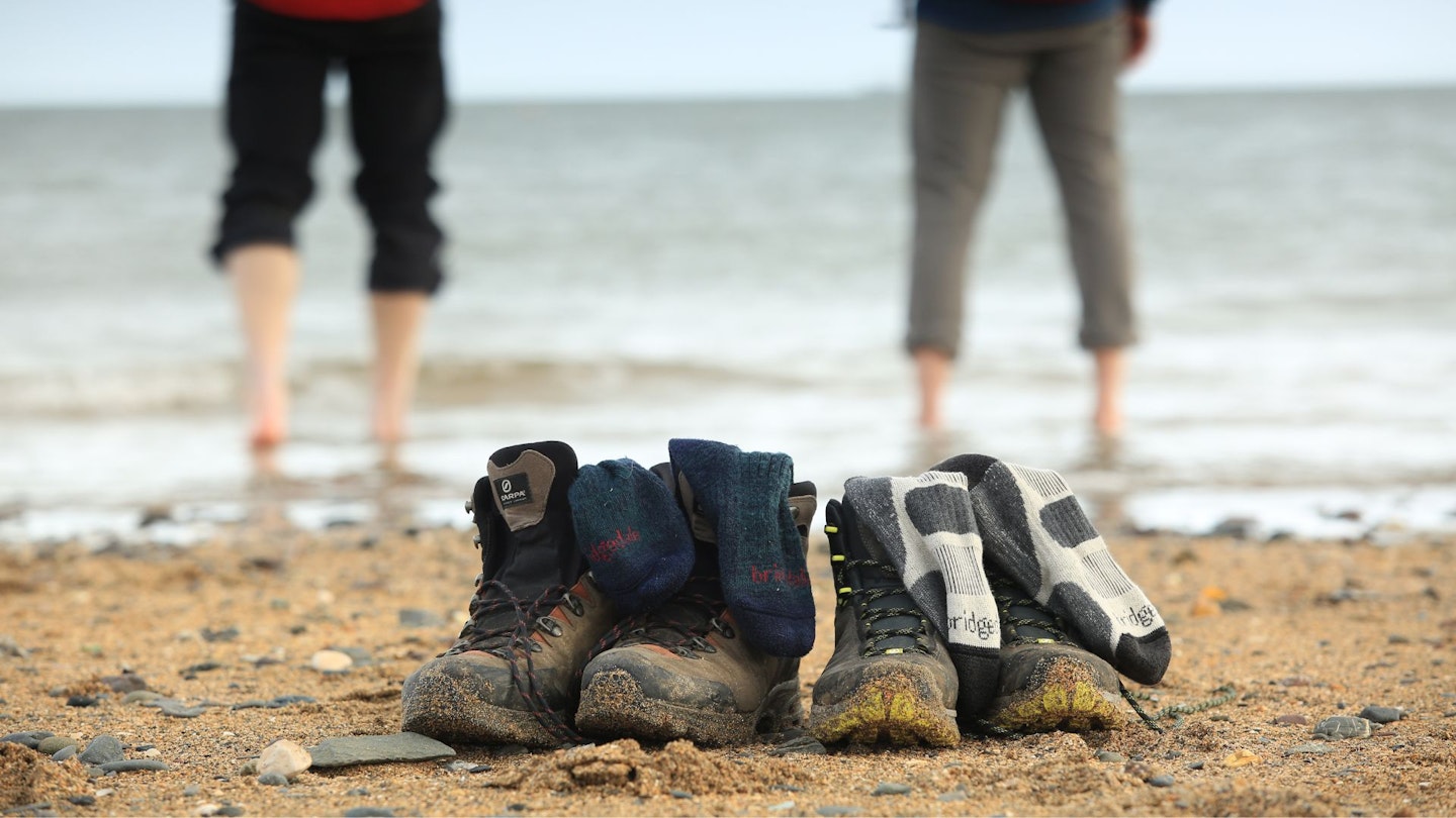 Two pairs of hiking boots with Bridgedale socks inside, with two hikers paddling in the sea in the background