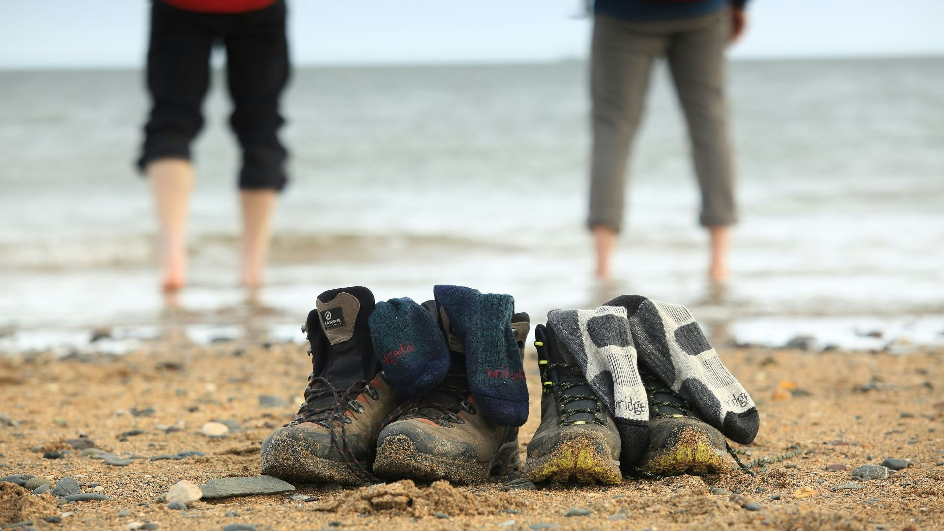Two pairs of hiking boots with Bridgedale socks inside, with two hikers paddling in the sea in the background