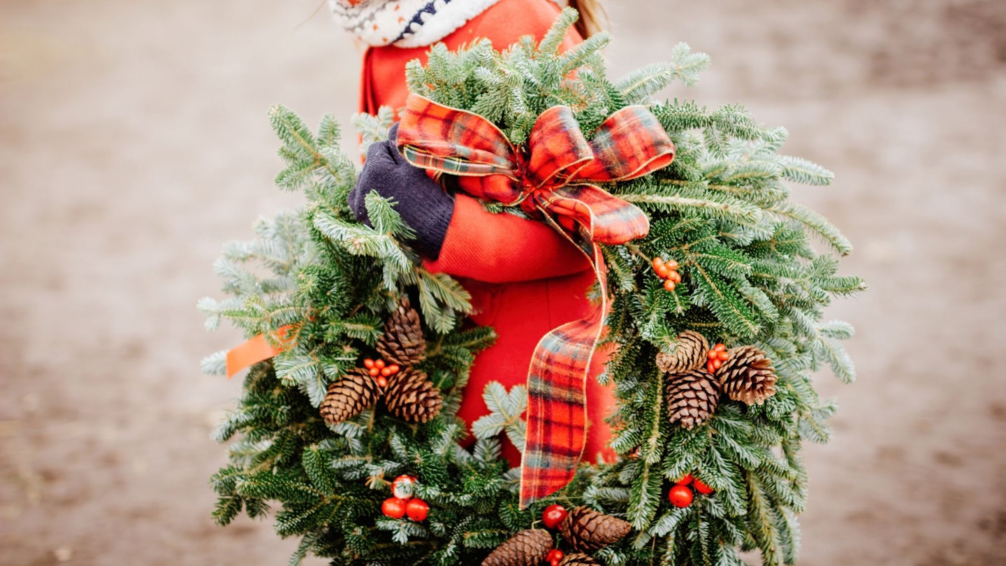 A girl carries a traditional Christmas wreath, that's decorated with red ribbons and pinecones