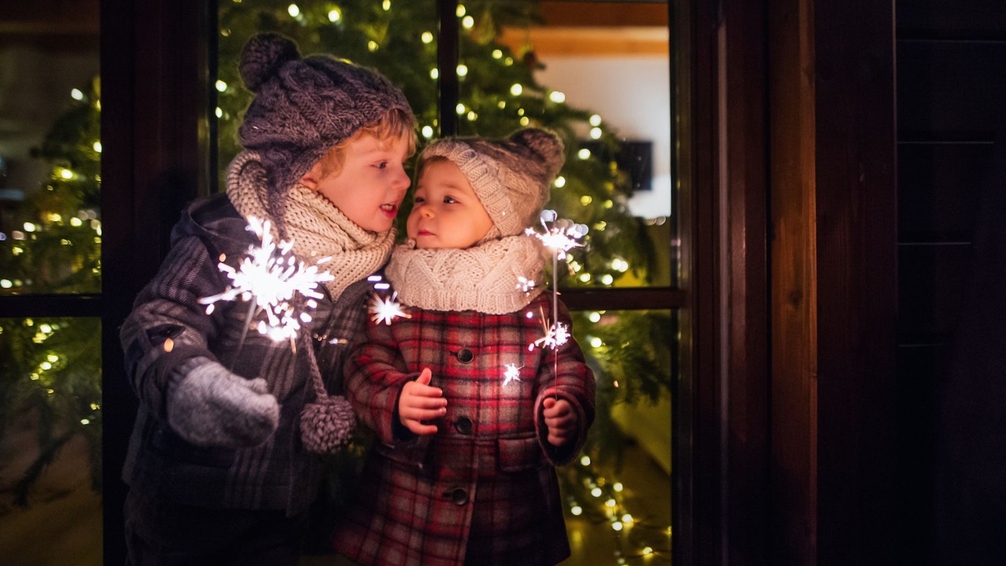 Two small children hold sparklers with a Christmas tree behind them.
