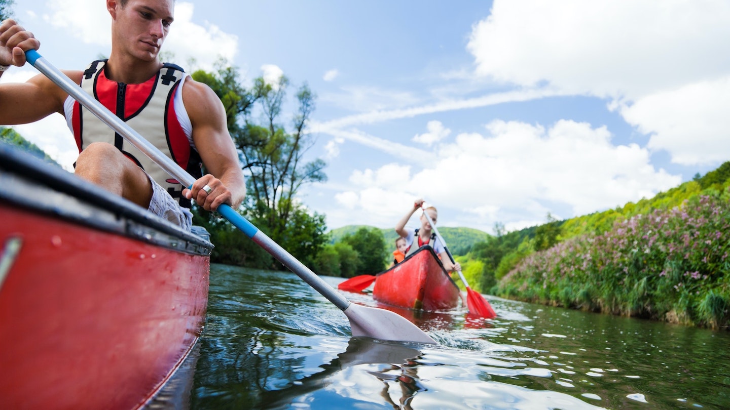 A man with strong arms paddles a kayak down a river, with people in the background