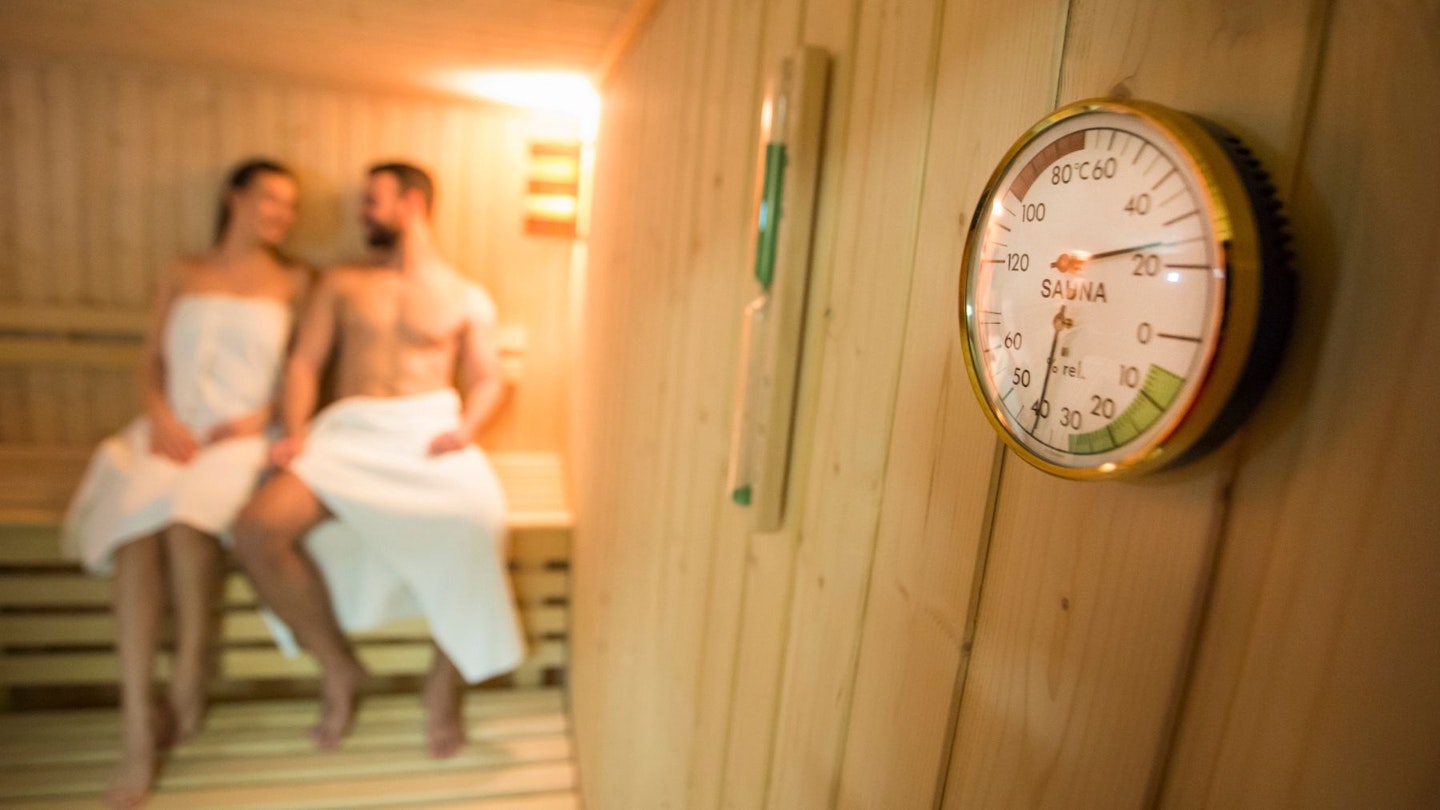 A couple sat in a sauna for the sauna health benefits, with the sauna dial in the fore screen.