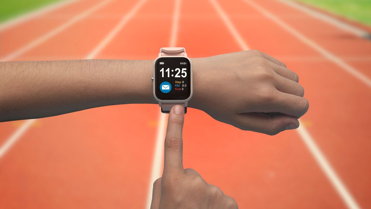 An individual pointing to a fitness smartwatch in front of a running track wearing the best wearable fitness tech